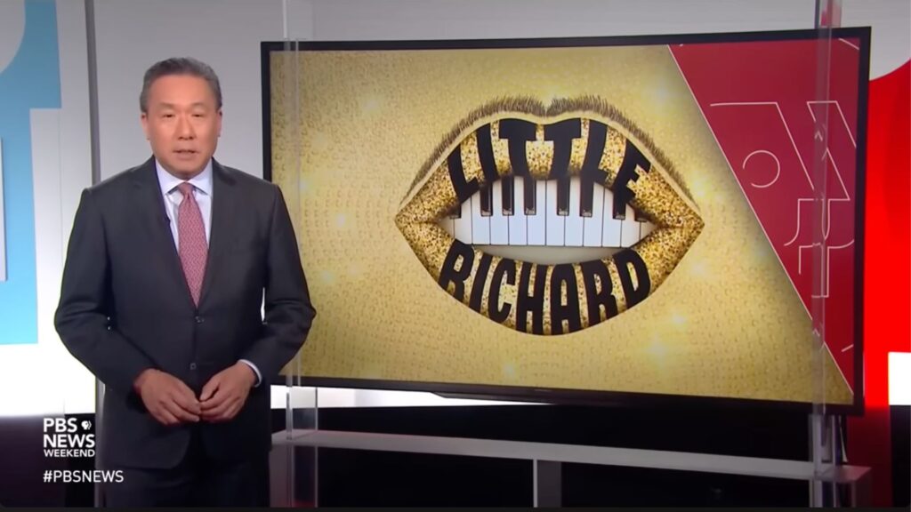 Cortés Filmworks: A news anchor from Cortés Filmworks stands in a studio next to a screen displaying an image of golden lips with piano keys for teeth and the words "Little Richard" written across them, highlighting his rock’n’roll legacy. The screen also features the PBS News Weekend logo in the lower left corner. : CortesFilmworks.com